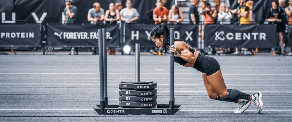 Femme poussant un traîneau lesté dans l'épreuve Sled Push du Hyrox. Test de puissance et de résistance musculaire.